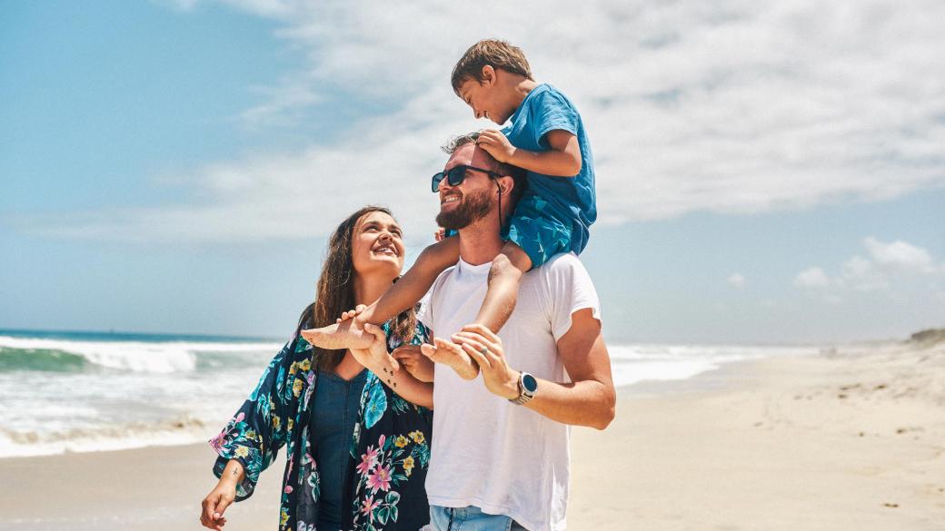 A smiling family walking along a summery beach with the father carrying his son on his shoulders