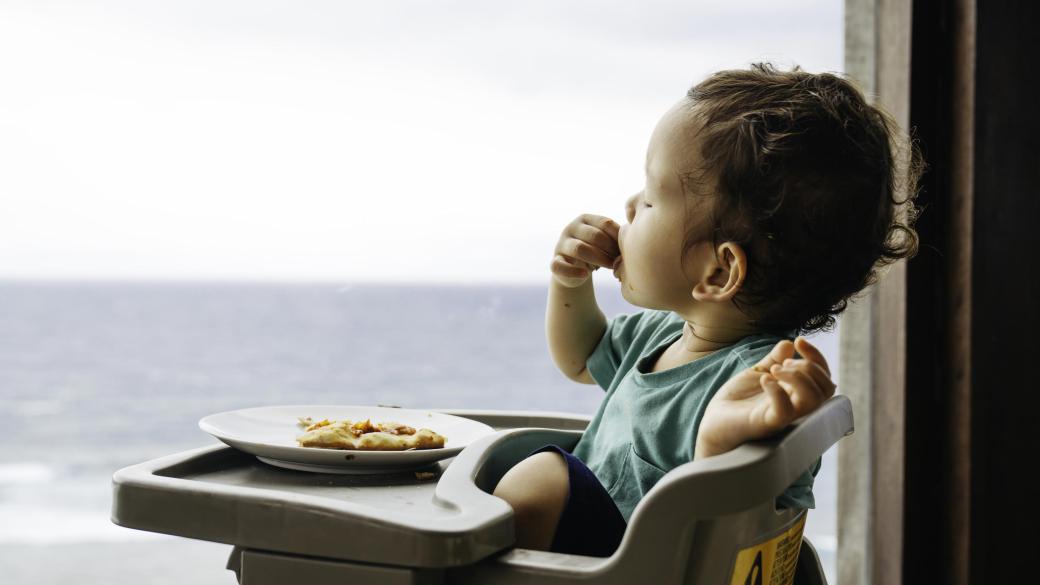 A toddler sitting in a high chair and eating a slice of pizza in front of a window with a view of the sea in the distance