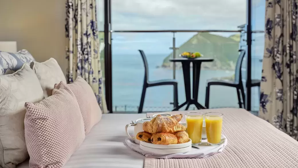 A breakfast tray with food and juice sitting on the bed in one of Sandy Cove Hotel's classic sea view with balcony rooms