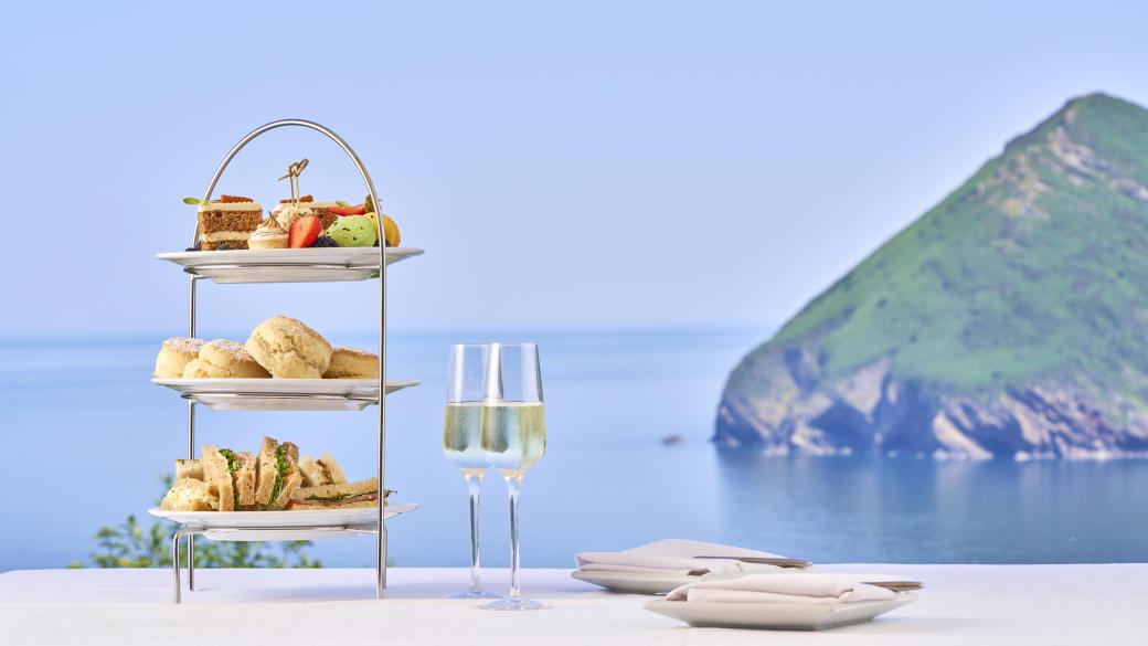 An afternoon tea cake stand with sandwiches, scones and cakes and two glasses of champagne in front of a view over the Bay