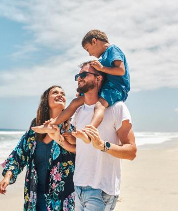 A smiling family walking along a summery beach with the father carrying his son on his shoulders