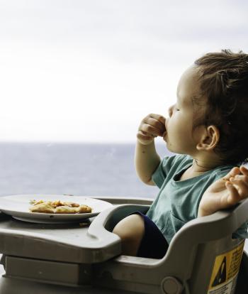 A toddler sitting in a high chair and eating a slice of pizza in front of a window with a view of the sea in the distance