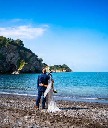 A bride and groom standing on Broadsands beach near Sandy Cove Hotel on their wedding day