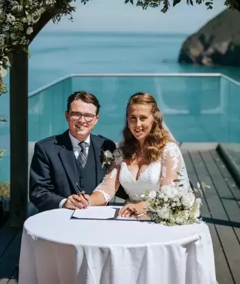 A smiling bride and groom signing their marriage licence on the platform outside The Venue at Sandy Cove Hotel