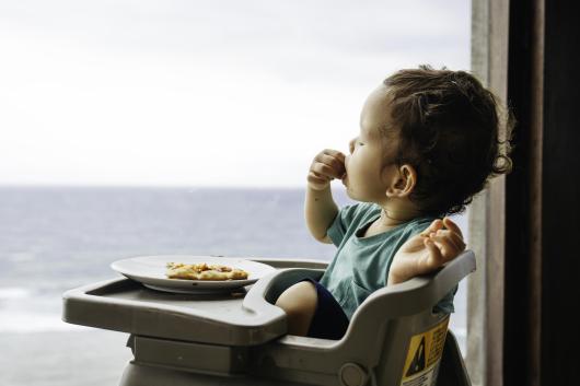 A toddler sitting in a high chair and eating a slice of pizza in front of a window with a view of the sea in the distance