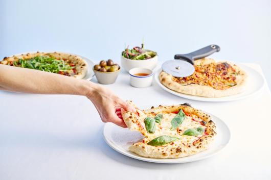 A hand taking a slice of Margherita pizza in The Cove Restaurant at Sandy Cove Hotel with other pizzas and side dishes in the background